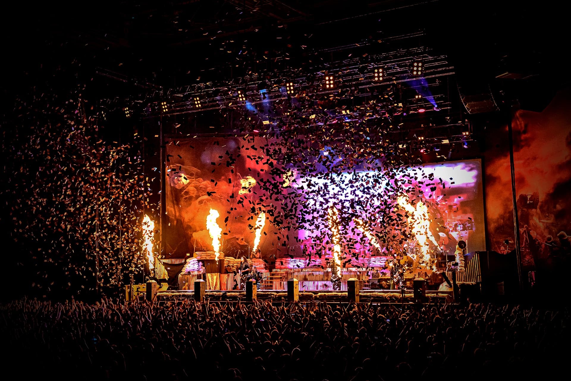 Main-stage concert with flame jets, confetti bursts and a packed crowd lit in red and magenta