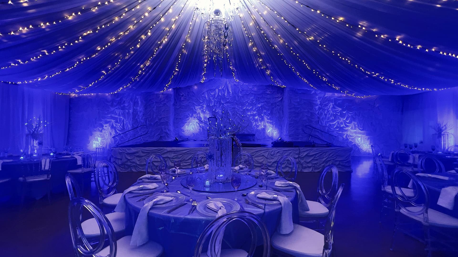 Blue-lit marquee with draped ceiling, fairy lights, chandelier and a set round banquet table with ghost chairs