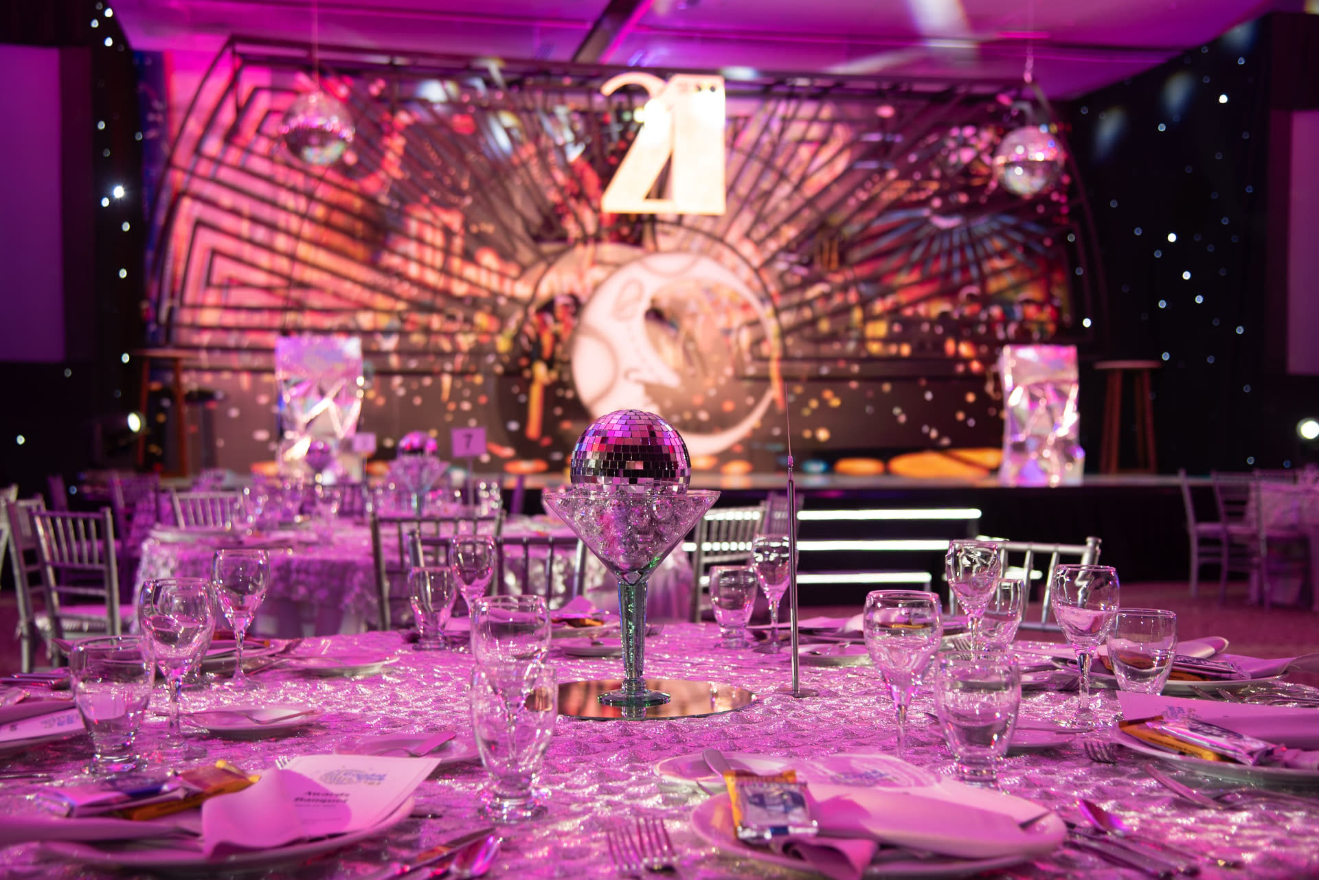 Magenta-lit gala banquet room with a mirror-ball centrepiece, set tables and a projected stage backdrop