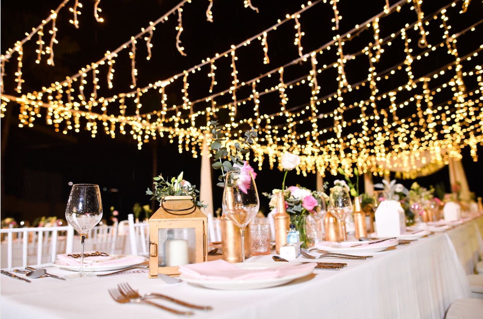 Warm fairy-light canopy above a long banquet table set with gold lanterns, florals and crystal glassware