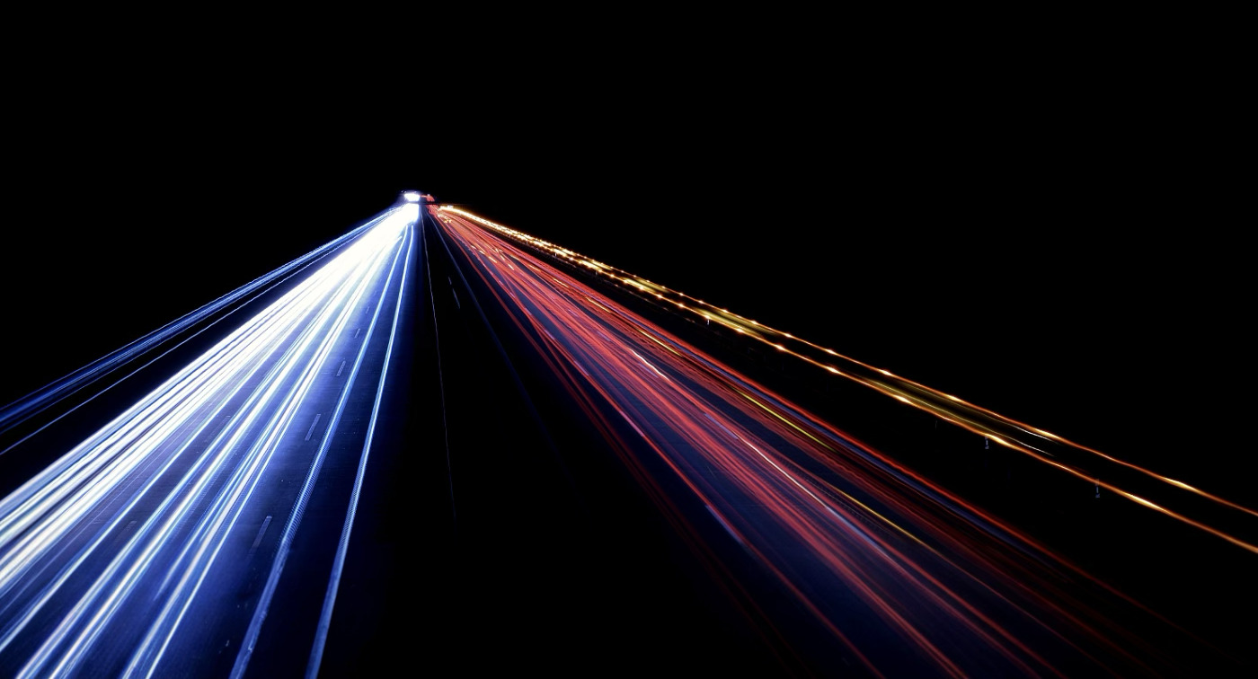 Long-exposure night highway with white, red and amber vehicle light trails converging to a single point on the horizon